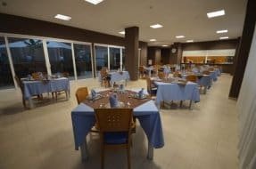 Comedor con mesas vestidas con manteles azules, sillas de madera y grandes ventanales con vistas al exterior.