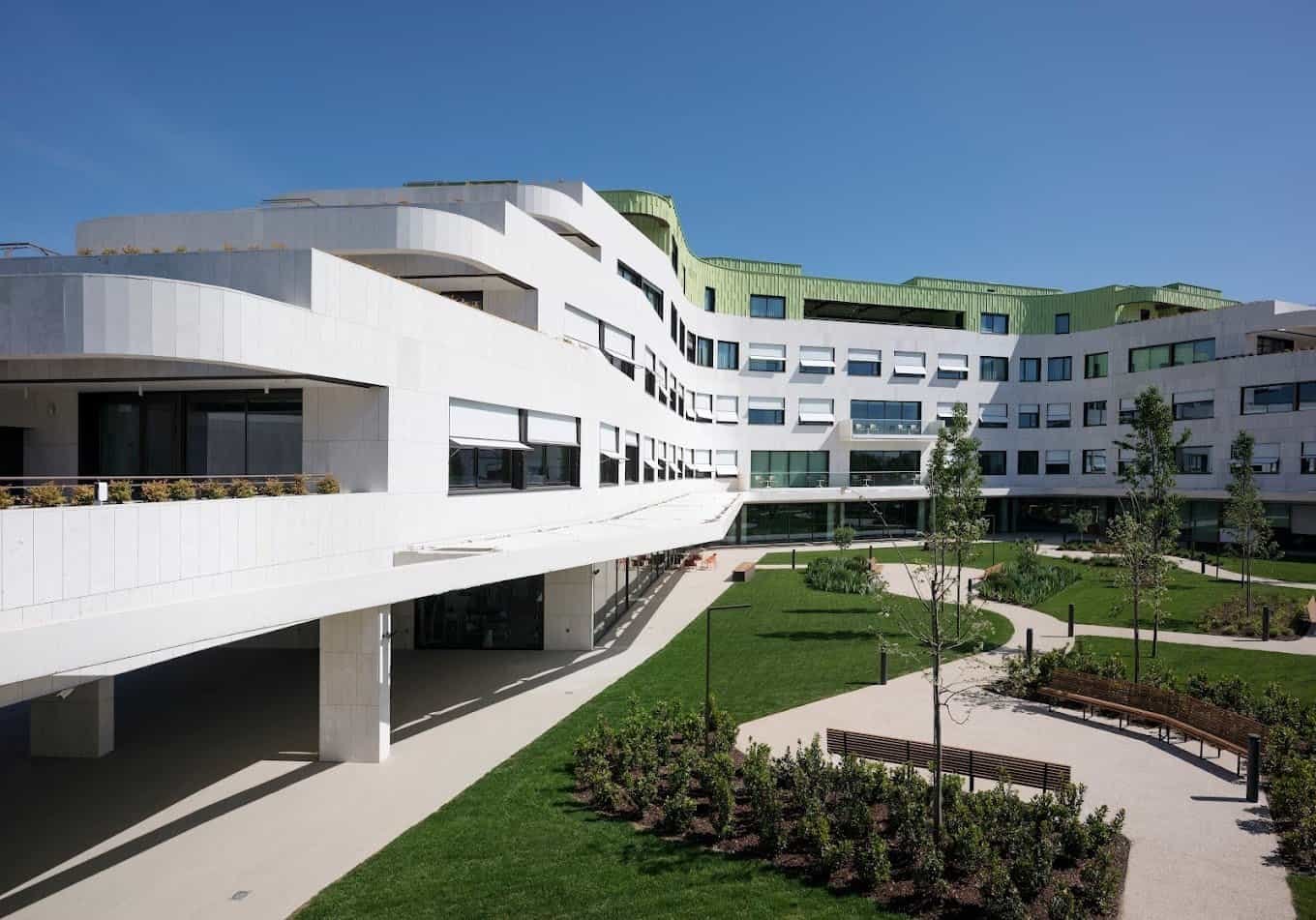 Edificio moderno de color blanco con un tejado verde y un jardín con caminos, césped y bancos.