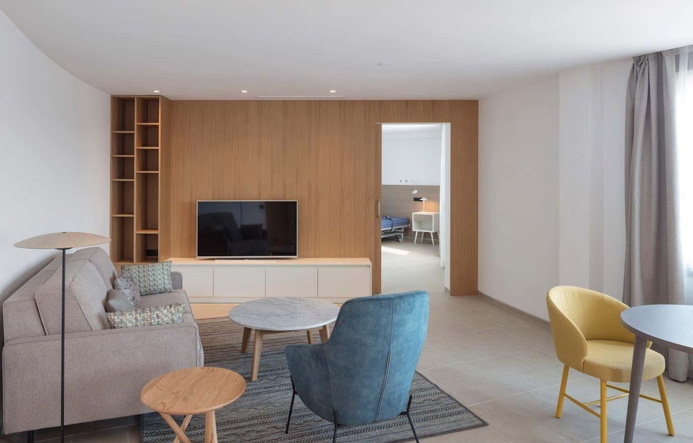 Sala de estar con un sofá, una televisión, una butaca de color azul y una butaca amarilla, con grandes ventanales.