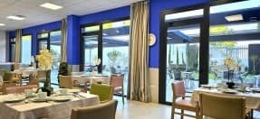 Comedor con mesas vestidas con manteles blancos, sillas de varios colores y grandes ventanales que dan a un patio.