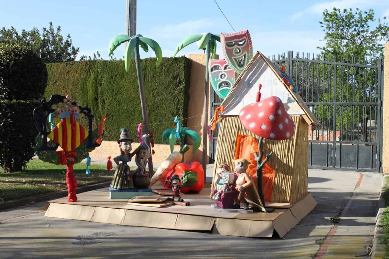 Escultura colorida con figuras de fantasía, palmeras y una casa de madera, en un patio exterior.