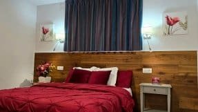 Habitación con una cama con colcha roja, un cabecero de madera, dos mesillas de noche y cuadros de flores en la pared.