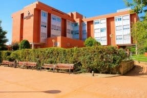 Fachada de un edificio de color beige con grandes ventanales, rodeado de un pequeño jardín con plantas.
