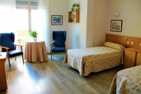 Habitación individual con una cama, un sillón de lectura, una mesilla de noche y una ventana con persianas.