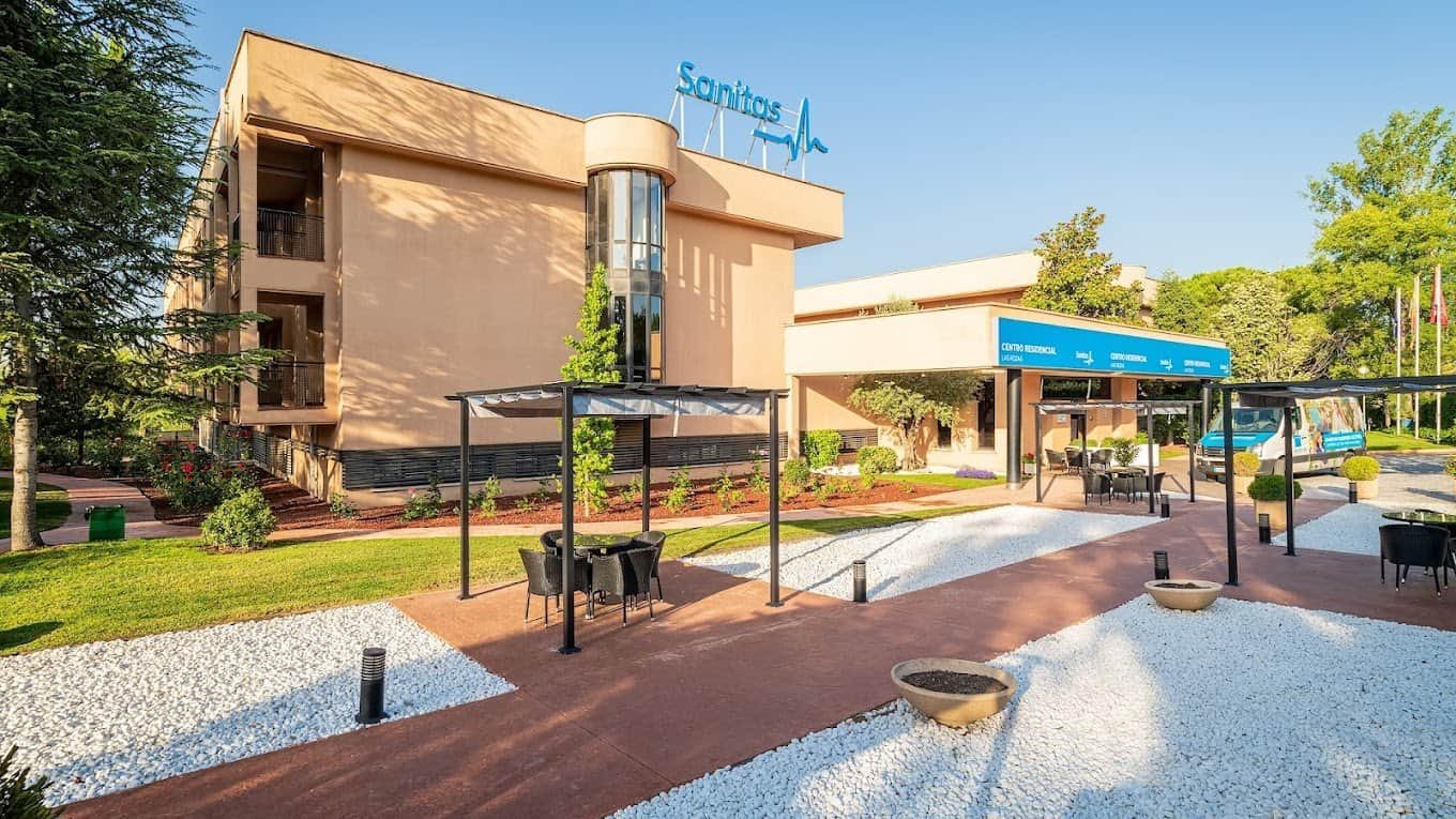 Exterior de un edificio de tonos ocres y marrones con el logotipo de Sanitas en la parte superior. El edificio tiene un área de jardín y una zona con mesas y sillas de exterior. Hay árboles y vegetación bien cuidada alrededor.