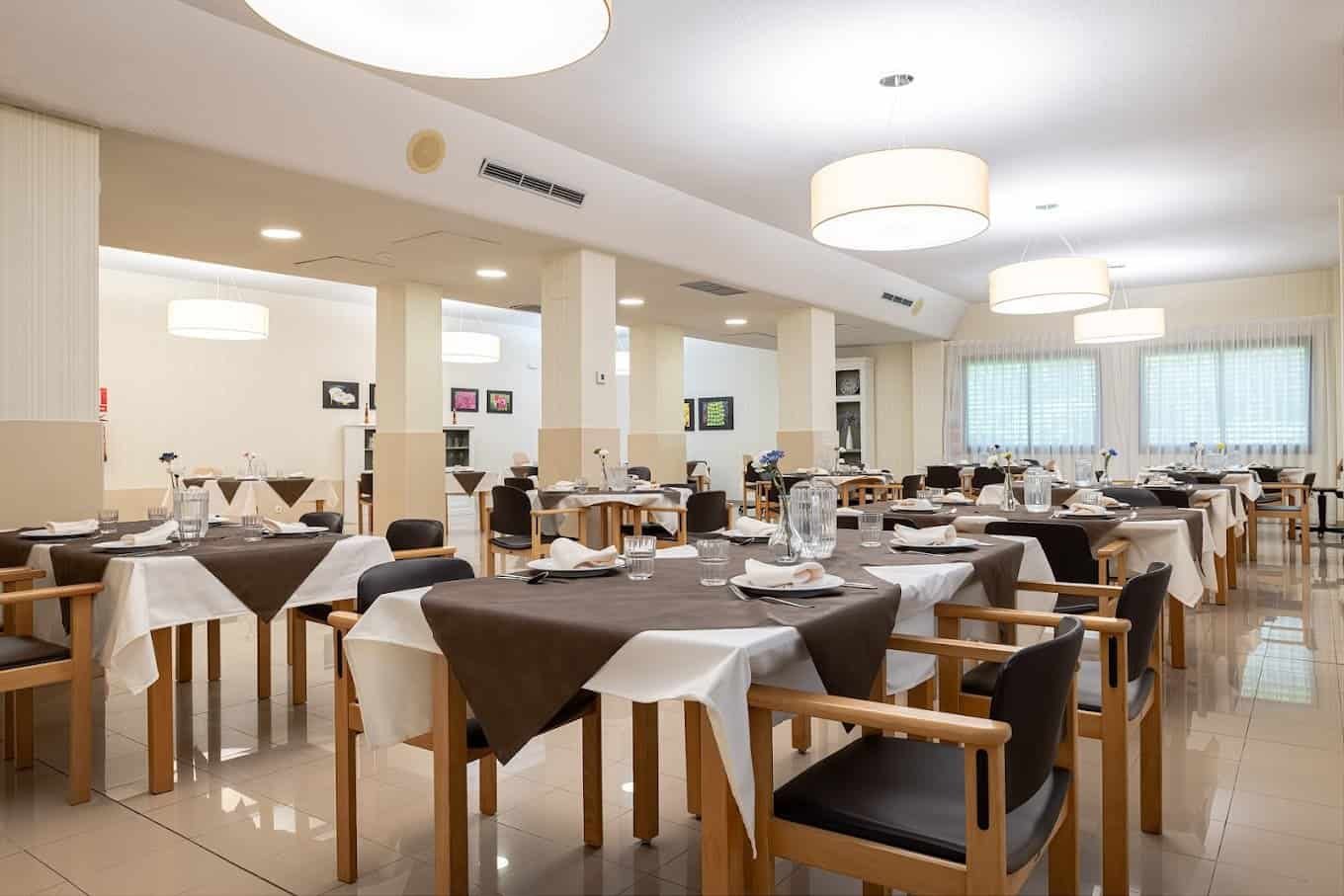 Vista interior de un comedor de restaurante con varias mesas redondas y cuadradas, cada una con manteles blancos y marrones, y sillas de madera oscura. El espacio está iluminado con varias lámparas de techo circulares y grandes ventanales.