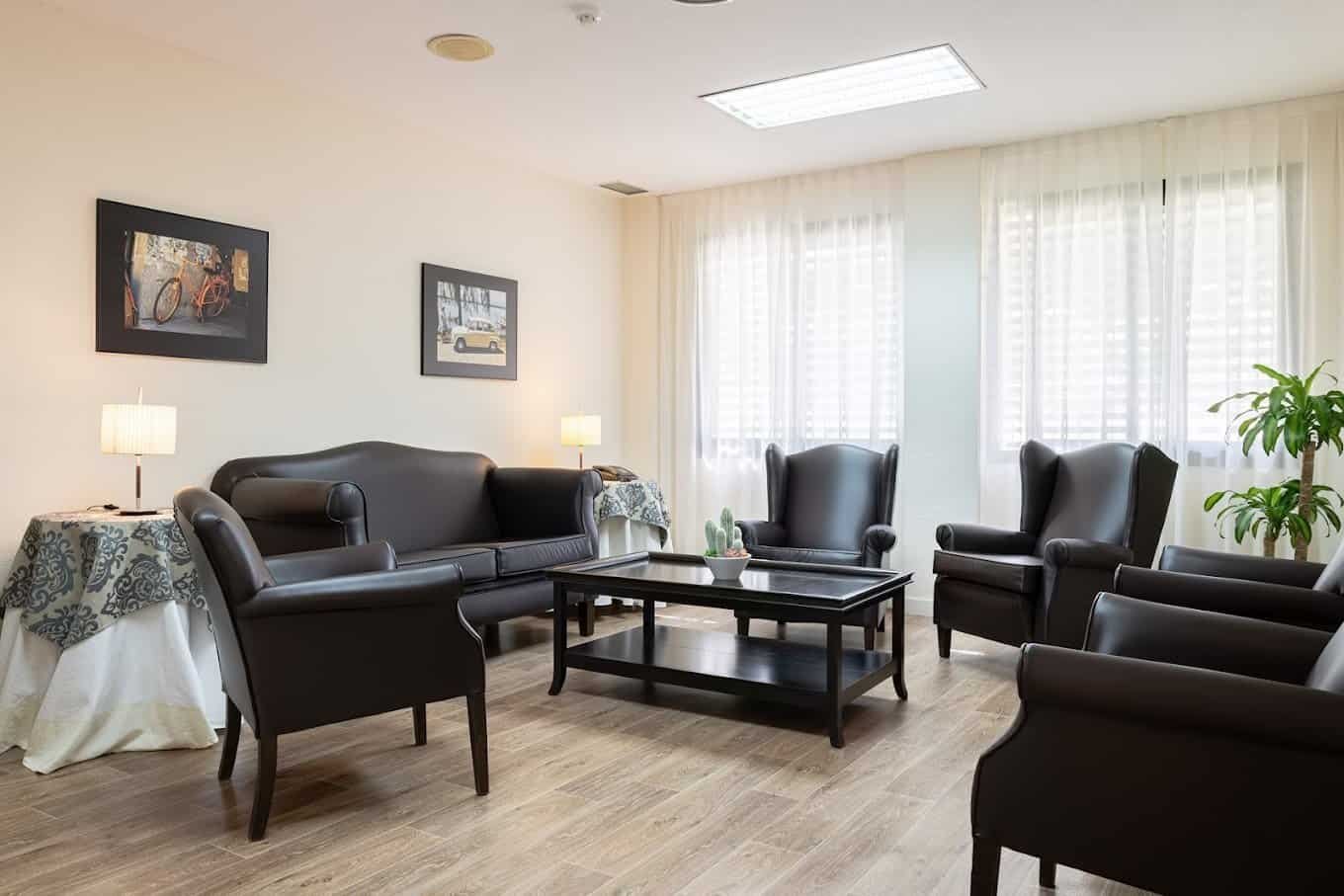 Interior de una sala de estar o área de espera con un sofá de cuero oscuro de tres plazas, un par de sillones individuales a juego y una mesa de centro de madera oscura. Hay dos mesas auxiliares con lámparas de mesa y cuadros decorativos en las paredes. El suelo es de madera clara y hay grandes cortinas blancas en las ventanas.