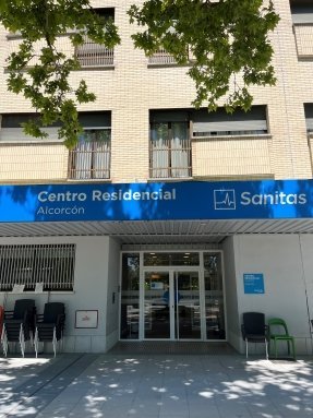Fachada exterior de Sanitas Residencia de Mayores Alcorcón, un edificio moderno con paneles de colores blanco y rojizo, y un gran jardín con césped.